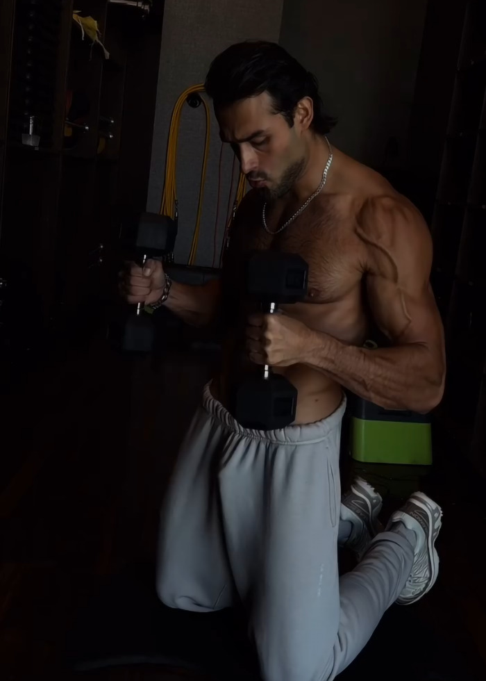 Sam Asghari shirtless and kneeling indoors lifting dumbbells during an intense workout session spotted by Britney Spears fans