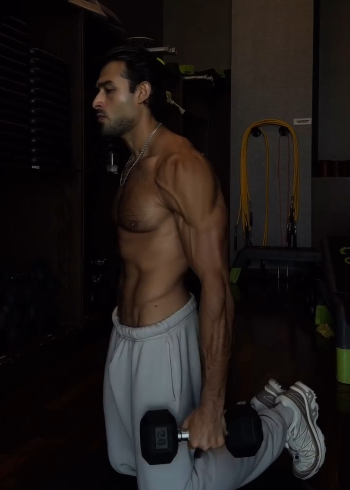 Sam Asghari shirtless in a gym, holding a dumbbell while exercising, showing muscular physique and intense focus.