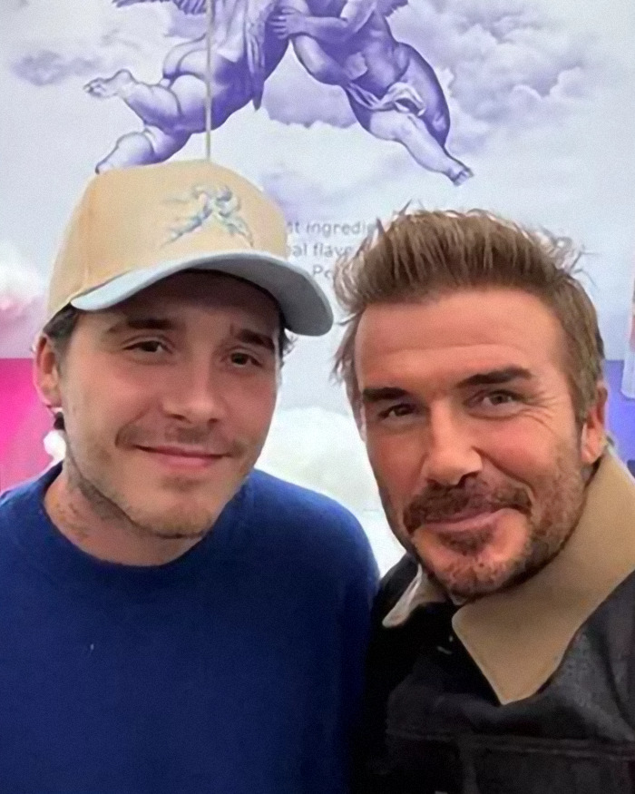 David Beckham smiling next to a young man in a cap with cherub artwork in the background during a casual moment.