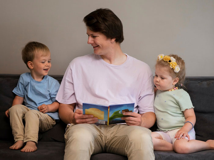 Man babysitting sister&rsquo;s kids on couch, reading a book, illustrating man agrees to babysit sister&rsquo;s kids trans narrative.