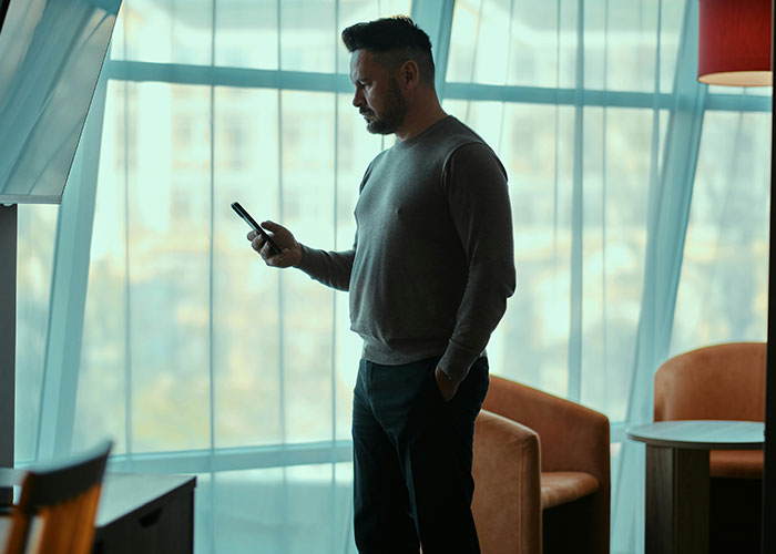 Man standing indoors by window, holding phone, reflecting on AITAH for telling brother's girlfriend about kids treatment boundaries.