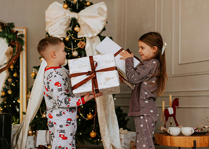 Two children in festive pajamas exchanging gifts near a decorated Christmas tree with wrapped presents around.