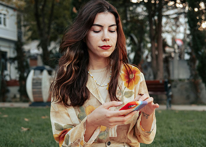 Young woman outdoors looking at phone, reflecting on telling brother's girlfriend about treating her kids differently.