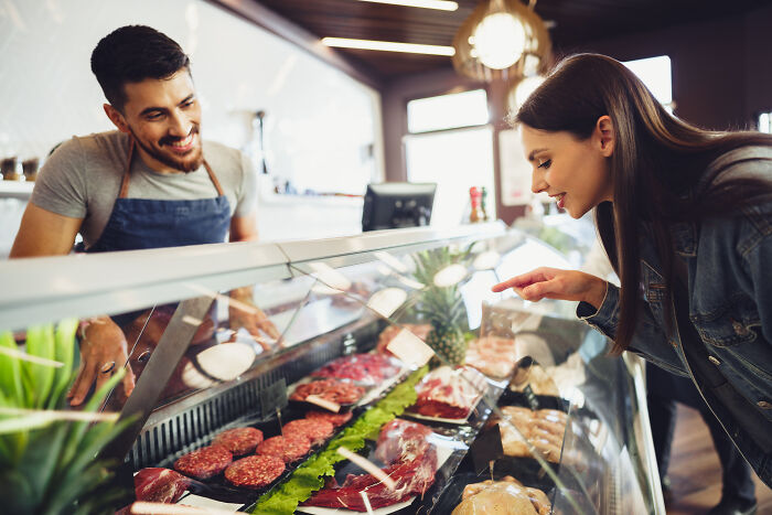 Man behind butcher counter smiling as female customer points at meat display, symbolizing company secret sharing.
