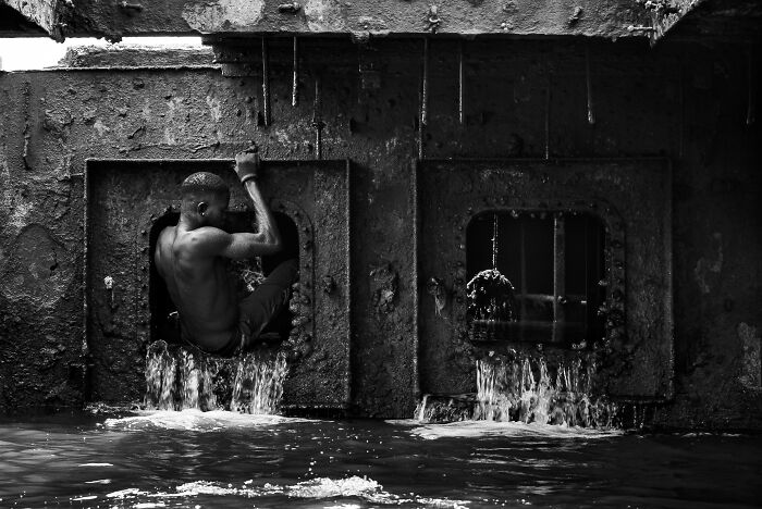 Young man climbing through a water-filled industrial structure, capturing the essence of humanity in a dramatic black and white image.