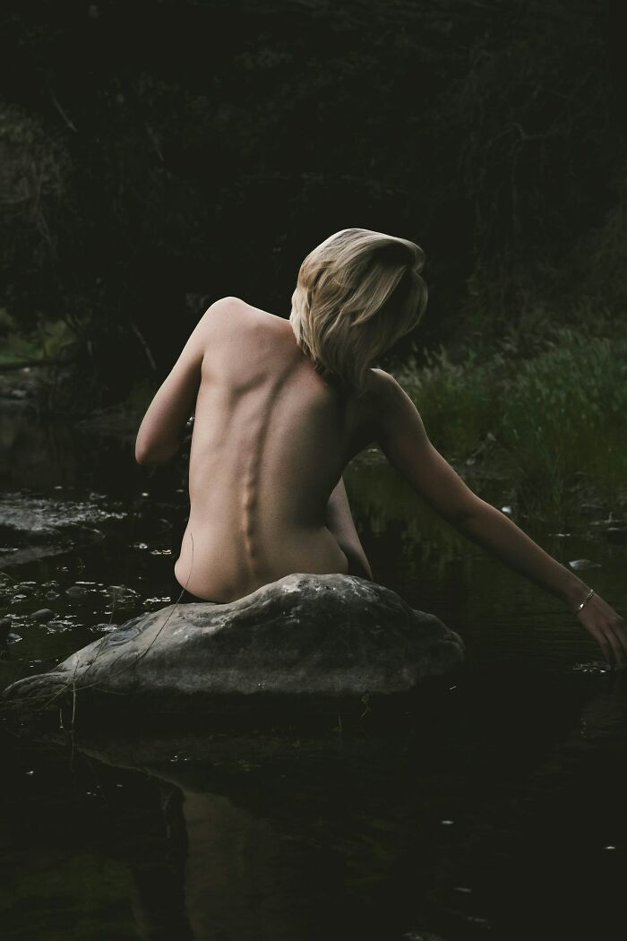 Person with bare back sitting on a rock near water in a dark natural setting, evoking weird and unhinged finds vibe.