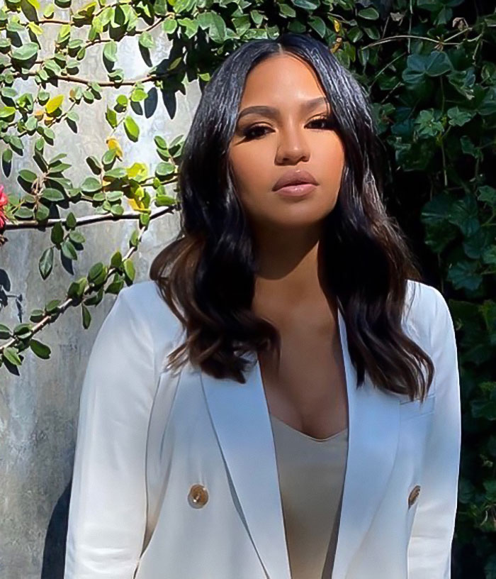 Cassie standing outdoors in a white blazer with greenery background, discussing freak-offs in a graphic detail setting.