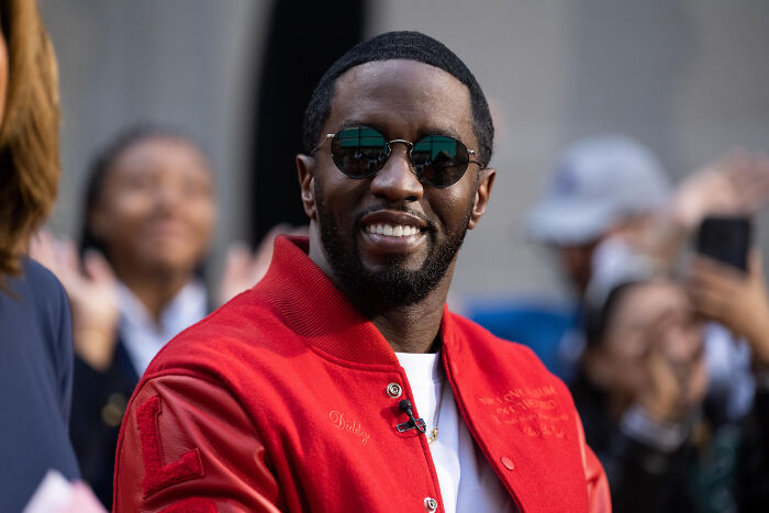 Man wearing sunglasses and a red jacket smiling at an outdoor event with blurred crowd in background, Diddy's ex-assistant.