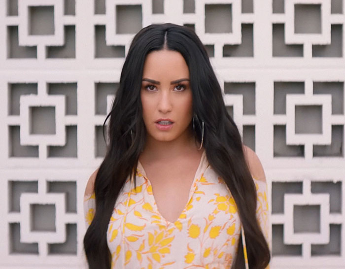 Young woman with long dark hair in a yellow floral dress posing in front of a decorative white wall, revealing celebrity insights.