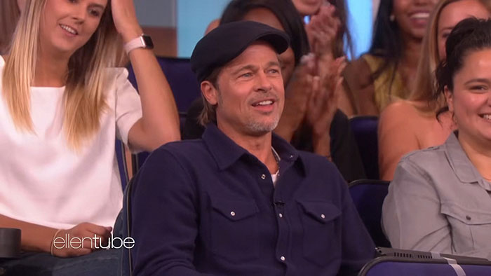 Brad Pitt wearing a navy jacket and cap, smiling during an interview surrounded by an audience on a TV show set.