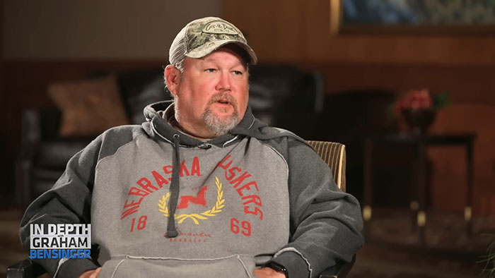 Middle-aged man in casual hoodie and cap, sharing stories about celebrities before their fame in a relaxed interview setting.
