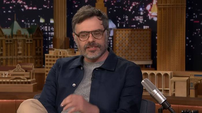 Actor wearing glasses and dark jacket sharing stories about celebrities before their fame during a late-night talk show interview.