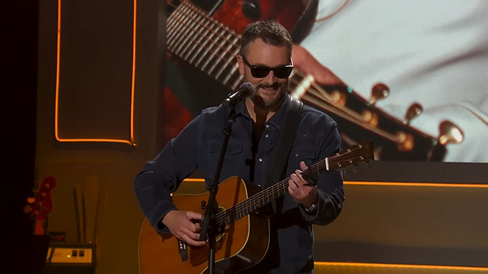 Male musician wearing sunglasses plays acoustic guitar and sings on stage with celebrity stories backdrop.