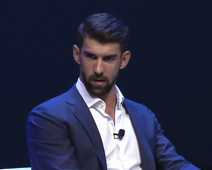 Man in a navy suit and white shirt speaking on stage, featured in stories about celebrities before their fame struck.