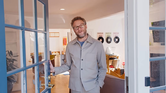 Man in glasses and casual jacket standing at an open door in a cozy living room, sharing celebrity insights before fame.