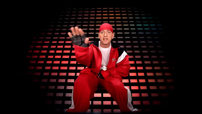 Eminem in a red tracksuit and bandana, performing on a stage with a red and black grid background.