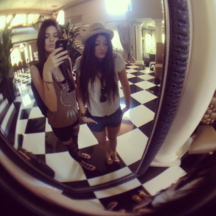 Two young women taking a mirror selfie indoors, showcasing casual vintage style in a checkered floor setting, celebrity posts.