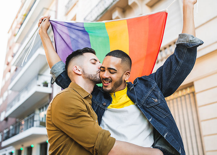 Two men embracing and smiling outdoors, one holding a rainbow pride flag, related to people who cheated on their partners.