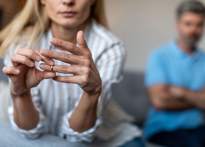 A woman removing her wedding ring with a distant partner in the background, symbolizing cheating confessions.