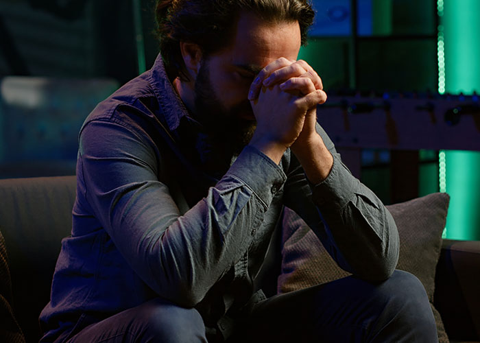 Man sitting on couch with head bowed and hands clasped, reflecting on cheating on their partner and emotional struggles.