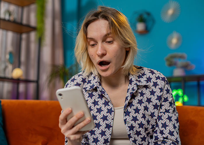 Young woman with shocked expression reading a confession about cheating on their partner on her phone indoors.