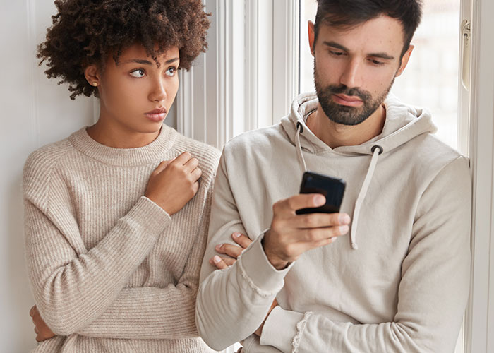 A tense couple stands close by a window, the woman looking worried and the man focused on his phone, illustrating cheating confessions.