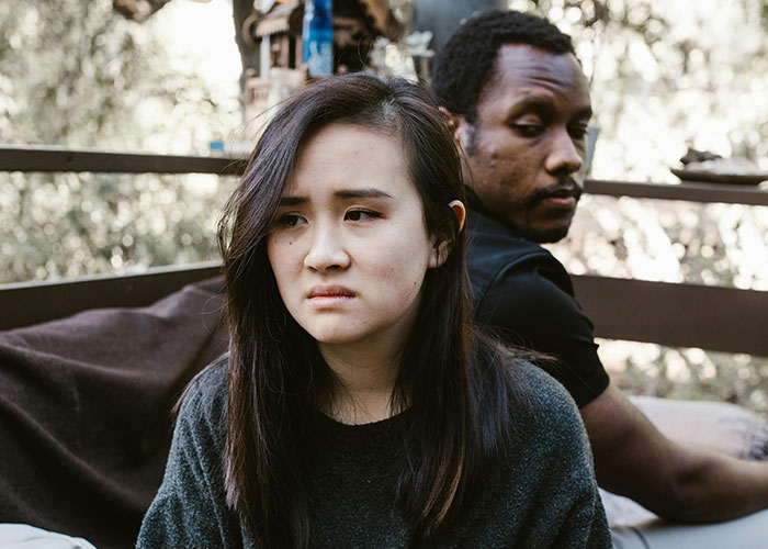 A conflicted couple sitting back to back outdoors, expressing emotions related to cheating on their partners.