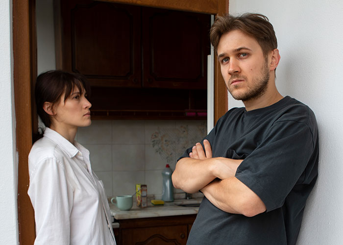 A tense young couple in a kitchen showing signs of cheating and relationship suspicion with crossed arms and serious expressions.