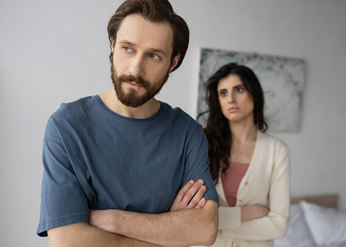 Man looking away with crossed arms while woman stands behind, both showing signs of relationship distrust and possible cheating.