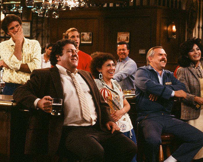 Cast of Cheers sitcom gathered at bar counter, smiling and laughing, highlighting fans heartbroken after Cheers star's passing news.