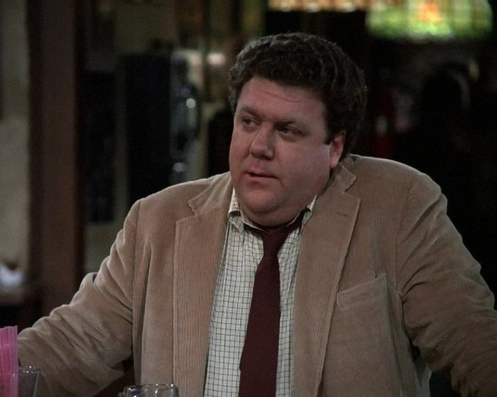 Actor John Goodman from Cheers wearing a beige jacket and red tie, appearing heartbroken after another Cheers star's passing.