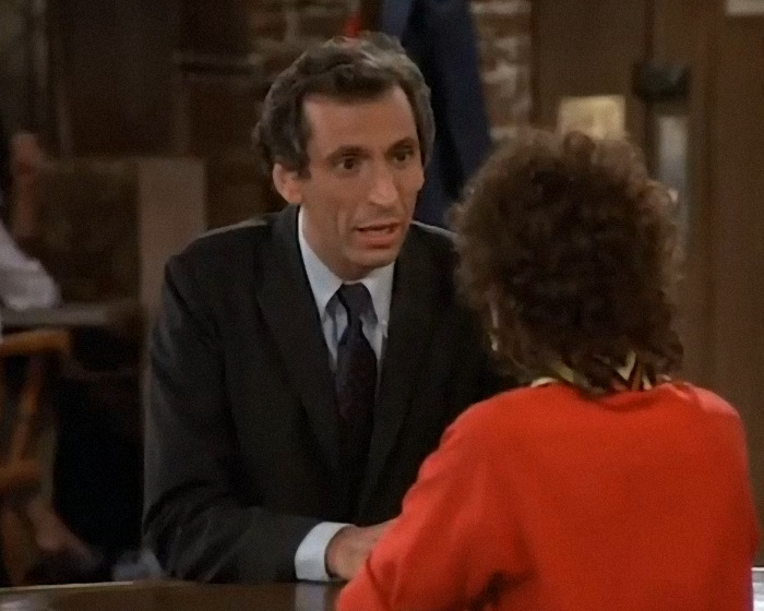 Cheers star in a dark suit talking seriously to a person in a red outfit inside a cozy bar setting.