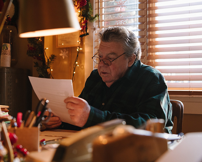 Older man with glasses reading a letter at a cluttered desk, expressing sadness about Cheers star's passing news.