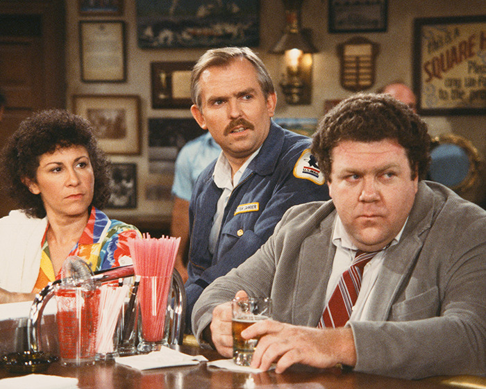 Three Cheers cast members sitting at a bar counter in a classic pub setting, reflecting on the show's legacy.
