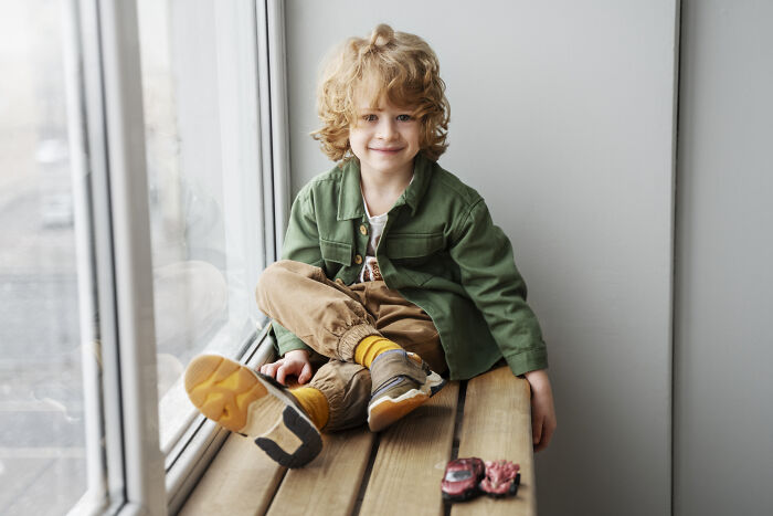Smiling young boy with curly hair sitting by a window wearing casual clothes illustrating hilariously bad names concept.