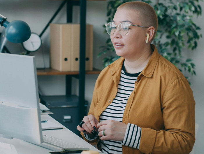 Person without kids working at a desk, wearing glasses and a mustard jacket, focused on a computer screen in an office setting.