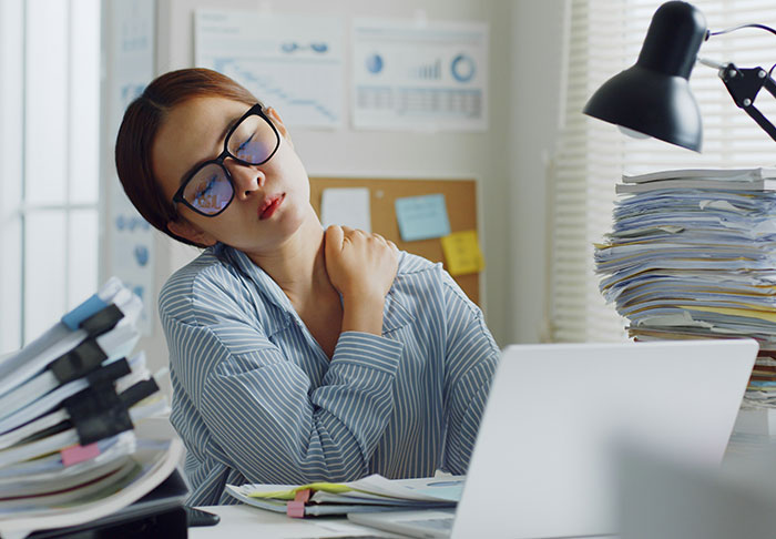 Tired woman in office rubbing her shoulder surrounded by paperwork, illustrating extra risky work situation without kids.