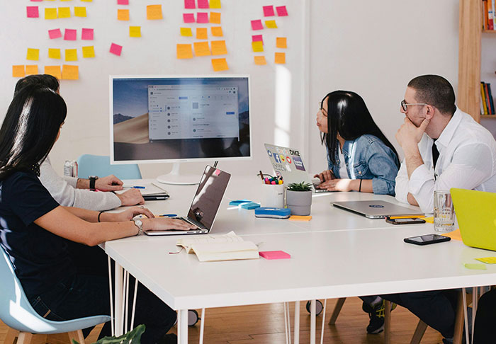 Four coworkers in a modern office with laptops and sticky notes, discussing a risky work nomination without kids.