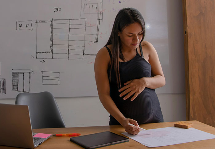 Pregnant woman working on a project at desk with laptop and whiteboard, illustrating extra risky work tasks without kids.
