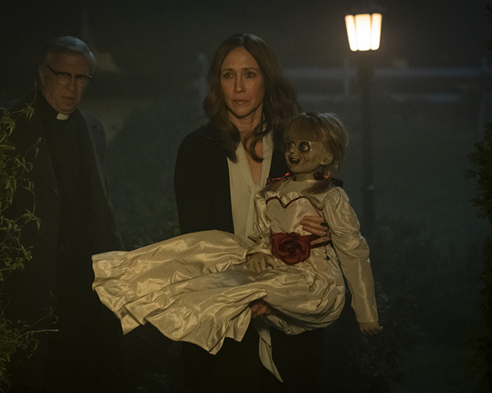 Woman holding haunted Annabelle doll at night with a priest in the background, highlighting the doll&rsquo;s chilling history.