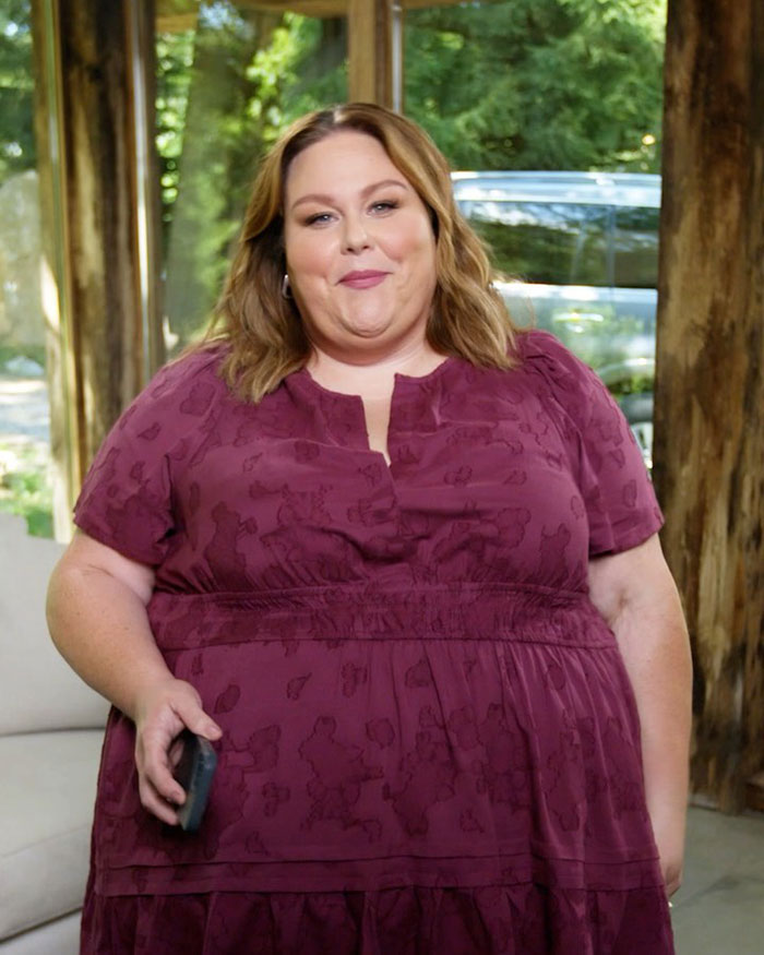 Chrissy Metz wearing a maroon dress, smiling indoors with nature visible through large windows behind her.