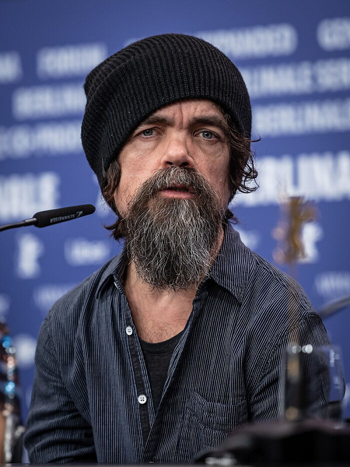 Man with a thick beard and black beanie speaking at a microphone, connected to celebrity gossip and juicy pieces shared.