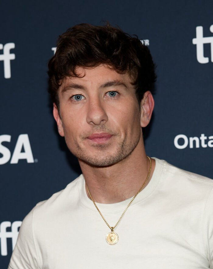 Young man with blue eyes and brown hair wearing a white shirt and gold necklace posing at a celebrity gossip event.