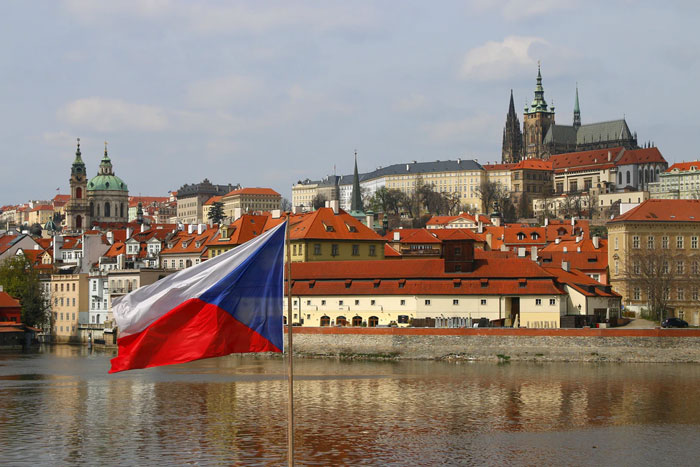 Czech Republic flag waving by the river with Prague cityscape reflecting classic national stereotypes humorously.