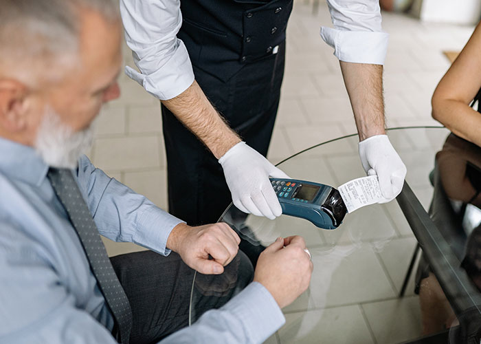 Man seated at glass table with waiter wearing white gloves handing over receipt and card machine, depicting national stereotypes.