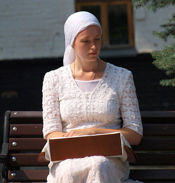 Woman in white headscarf and dress sitting on a bench holding a box, reflecting a national stereotypes theme.