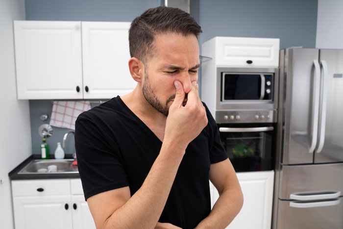 Man in kitchen holding nose with disgusted expression, reacting to cat-food-flavored coffee after mug theft.