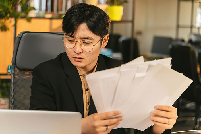 Man in glasses looking stressed while holding papers at office desk, depicting helicopter mom calls to son's boss scenario.