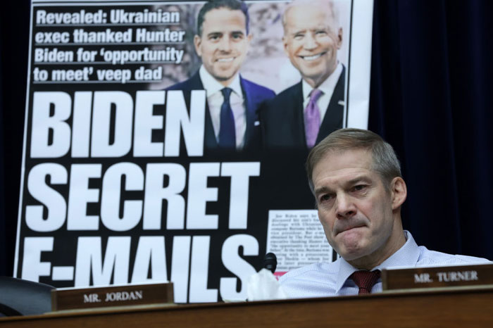Man sitting in front of a large screen displaying conspiracy theory headlines about Biden secret emails and Hunter Biden.