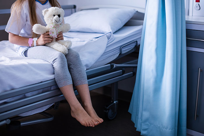 Child patient sitting on a hospital bed holding a teddy bear after attempting DIY medical treatment with injuries visible.